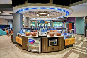 A modern hotel lobby with a central bar area featuring a refrigerated display, a coffee machine, and a seating area with blue chairs. - Tru by Hilton Sterling Heights Detroit (Sterling Heights)
