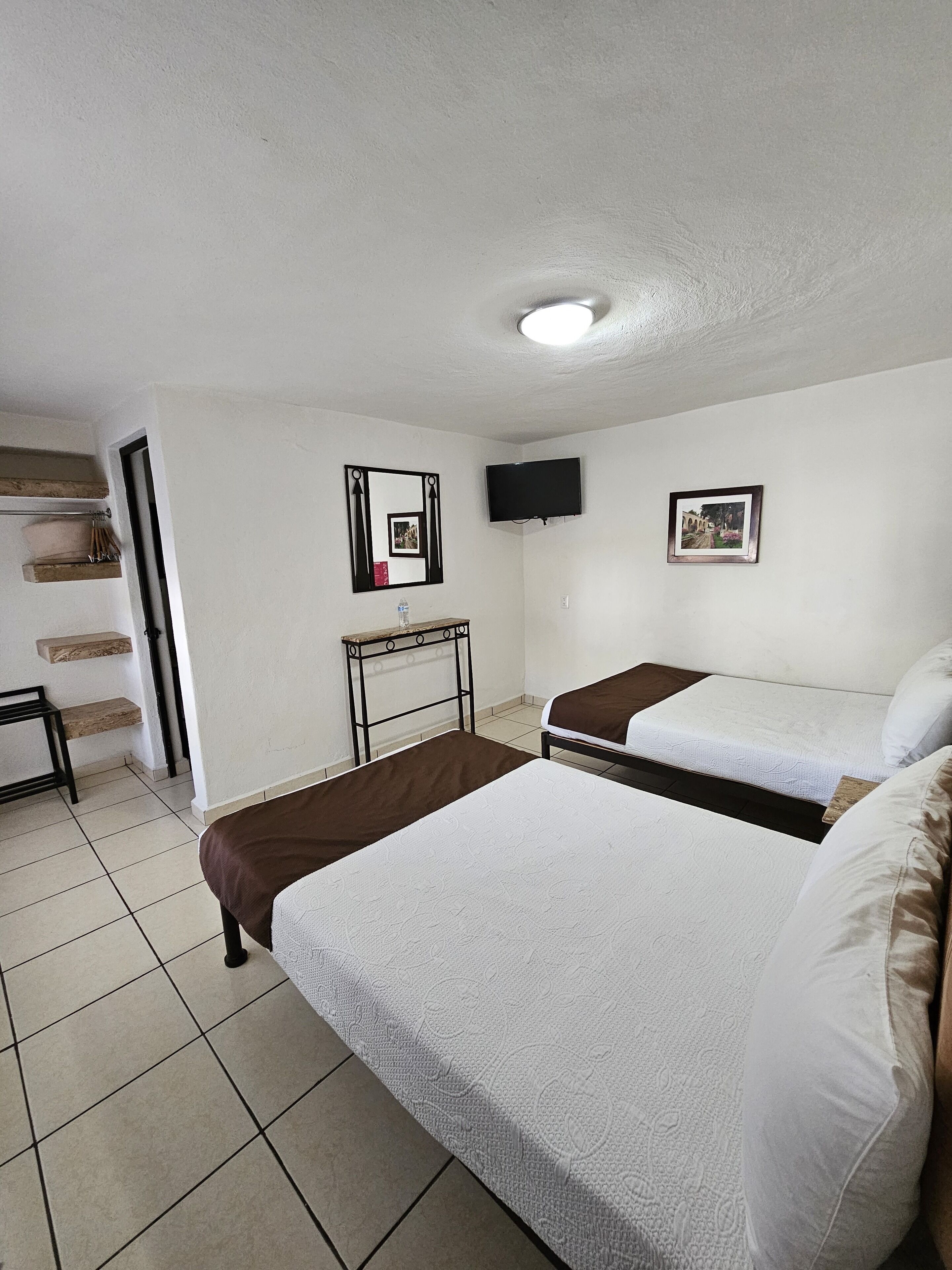 A hotel room with two beds, a TV mounted on the wall, a small table, and a shelf with items.