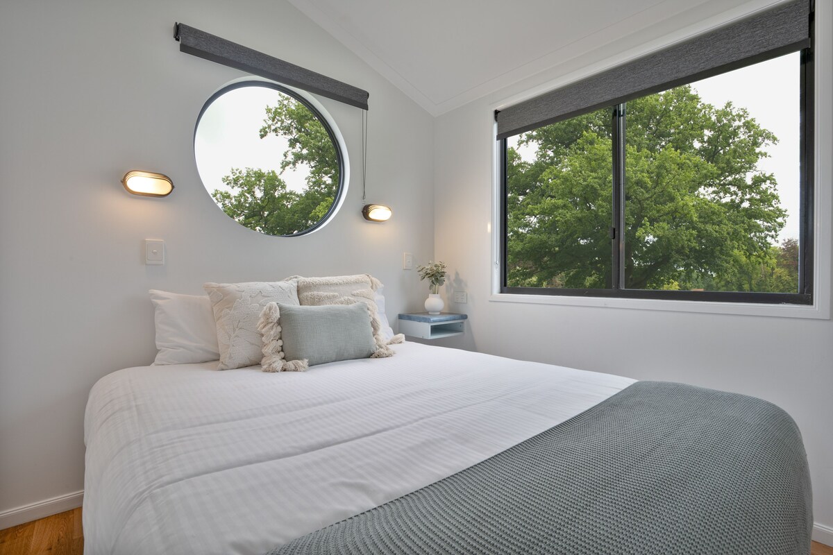 Een slaapkamer met een groot bed, een ronde spiegel, bedlampjes en een raam met uitzicht op bomen.