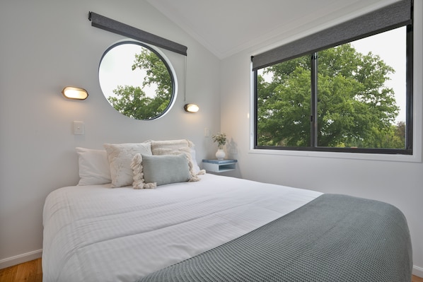A bedroom with a large bed, a round mirror, bedside lamps, and a window with a view of trees.