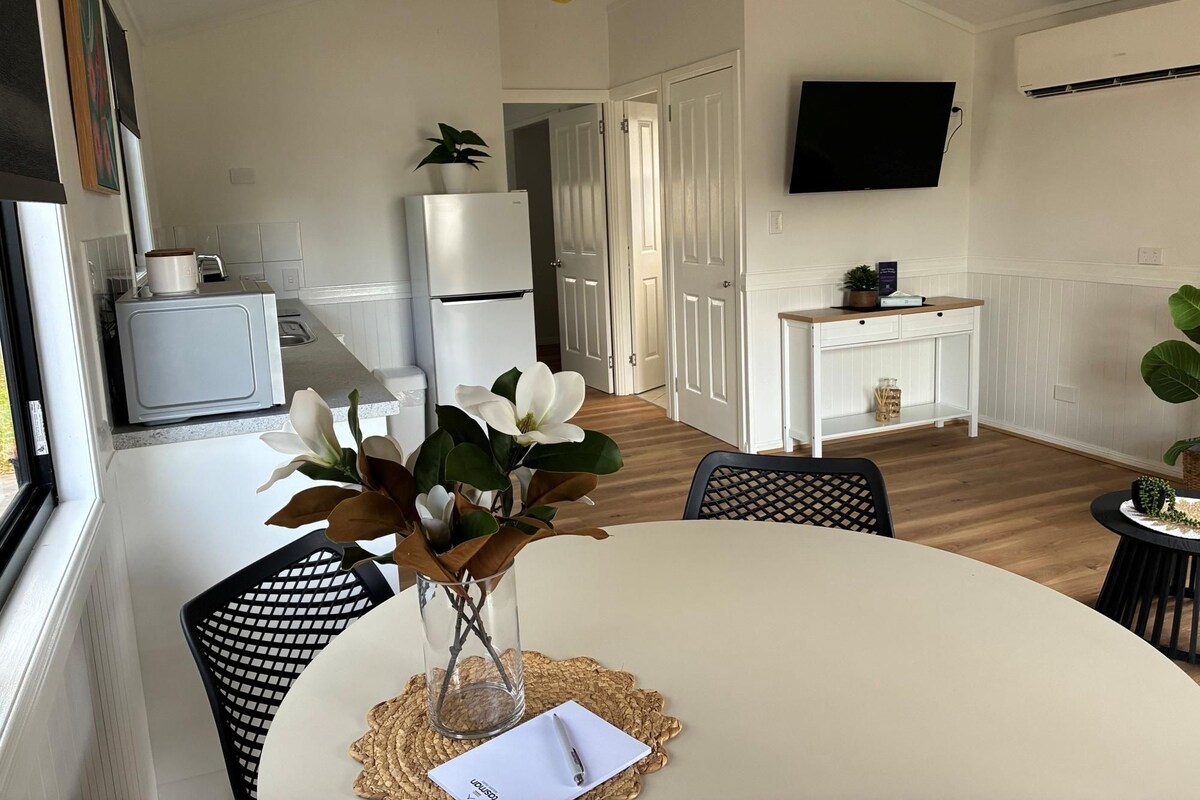 Een moderne keuken met witte apparaten, een eethoek met een witte tafel en stoelen, en een flatscreen televisie aan de muur.
