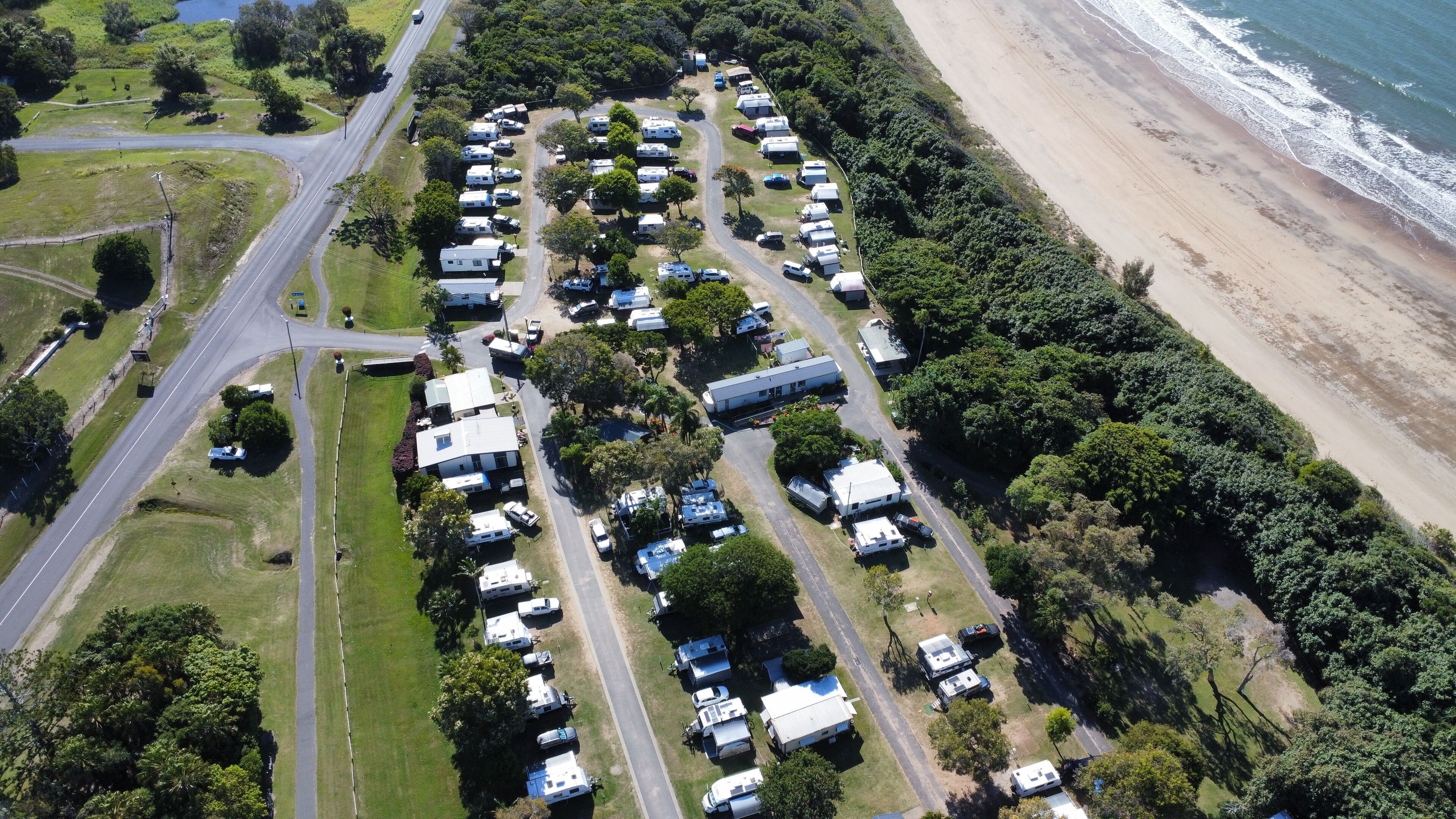 An aerial view of a campground with RVs and trailers parked along a road, adjacent to a beach.