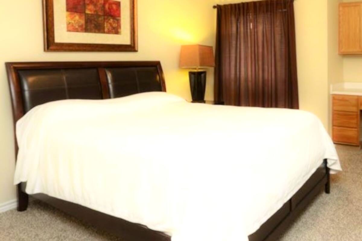 A neatly made bed with a white comforter in a room with a dark headboard, a framed picture on the wall, a lamp, and a wooden cabinet.
