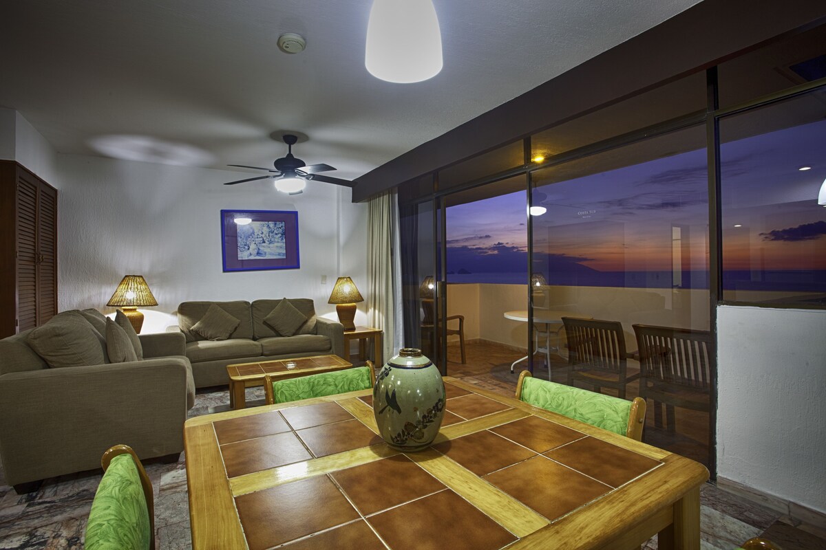 Una sala de estar moderna con un sofá, una mesa y un florero. Hay un ventilador de techo y una vista del atardecer a través de ventanas amplias.