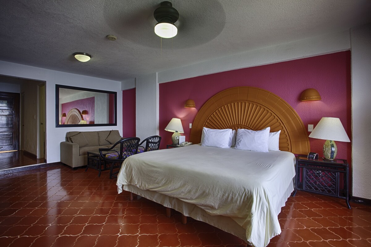 Habitación de hotel con una cama grande, un sofá, dos sillones, una mesita de noche con lámparas y un ventilador de techo.