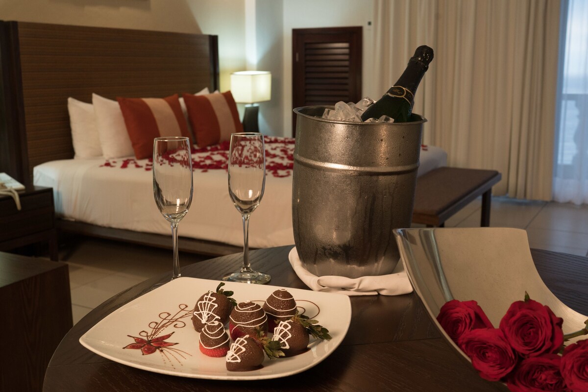 Habitación de hotel con cama, mesitas de noche, lámparas de noche, teléfono, una fuente de fresas bañadas en chocolate, dos copas de champán y una botella de champán en un cubo, junto con un ramo de rosas rojas.