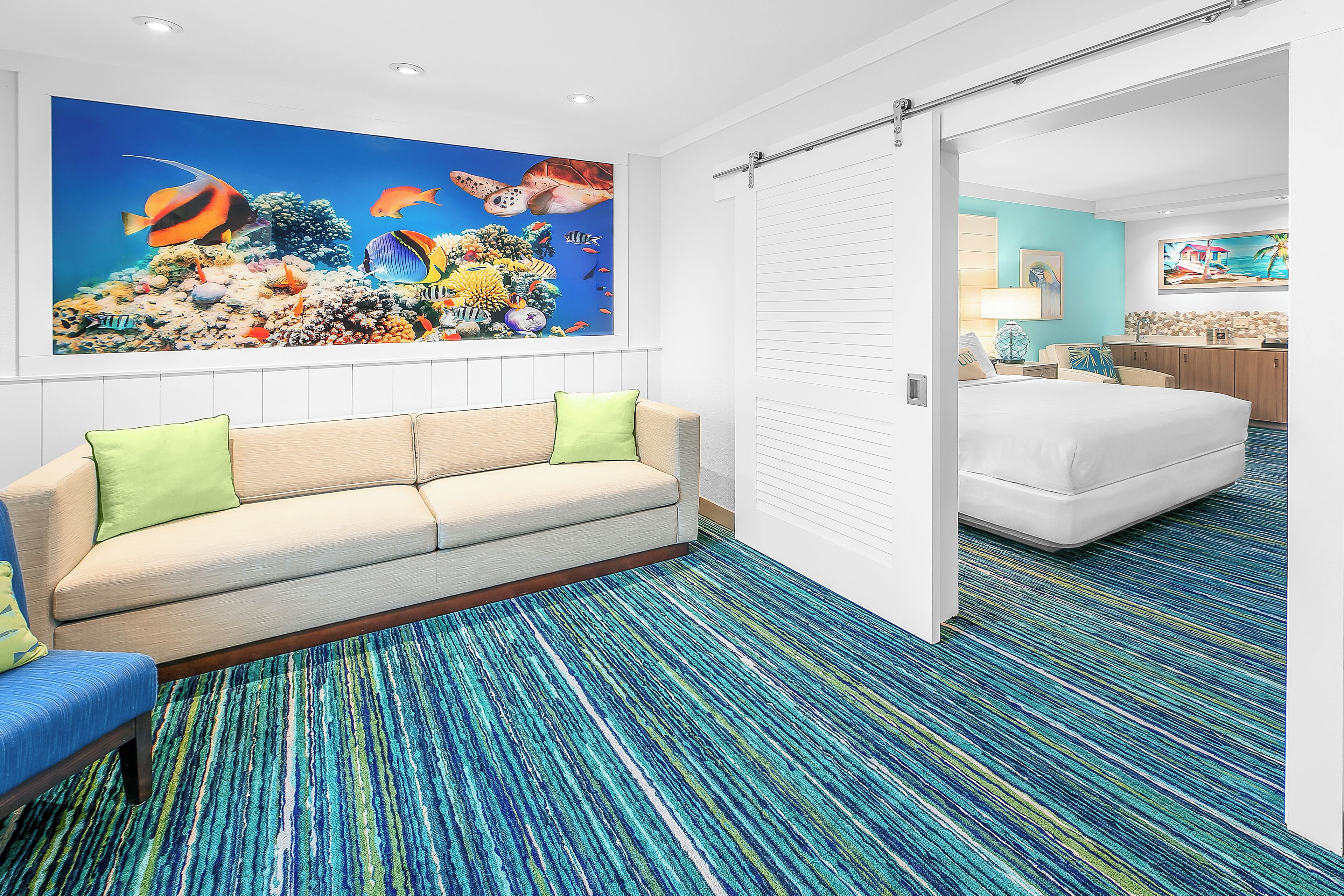A hotel room with a large ocean-themed mural, a sofa with green cushions, a patterned carpet, and a bed with a white comforter.