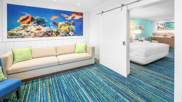 A hotel room with a large ocean-themed mural, a sofa with green cushions, a patterned carpet, and a bed with a white comforter.