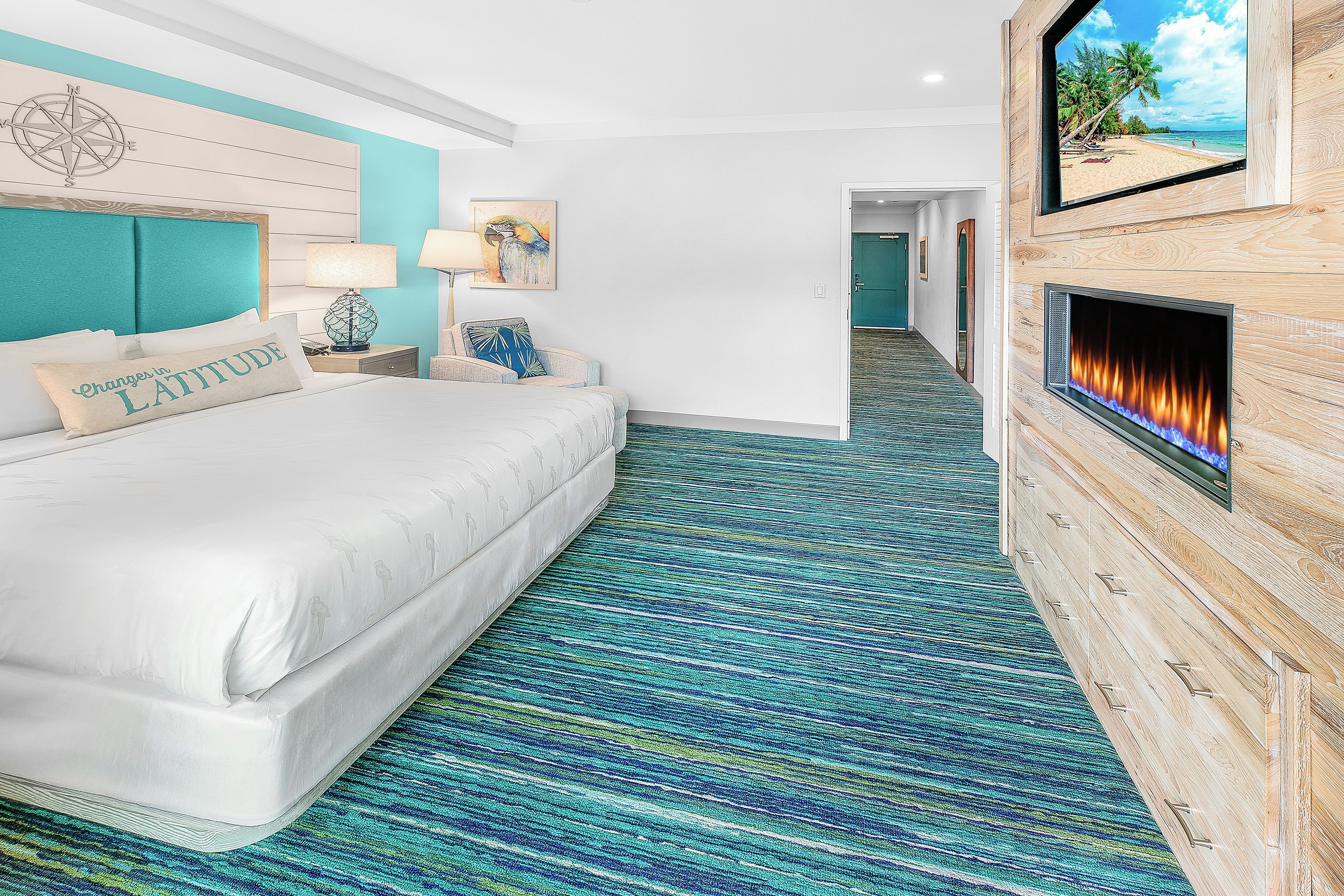A hotel room with a bed, a wall-mounted TV, a fireplace, and a view of a beach through a window.