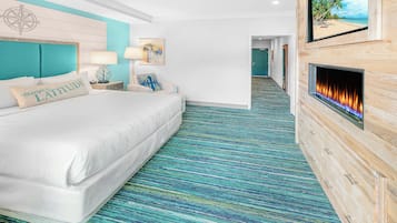 A hotel room with a bed, a wall-mounted TV, a fireplace, and a view of a beach through a window.
