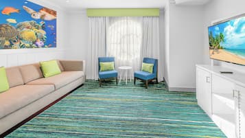 Habitación de hotel con un mural colorido, un sofá, dos sillones, una mesita y un televisor que muestra una escena de playa.