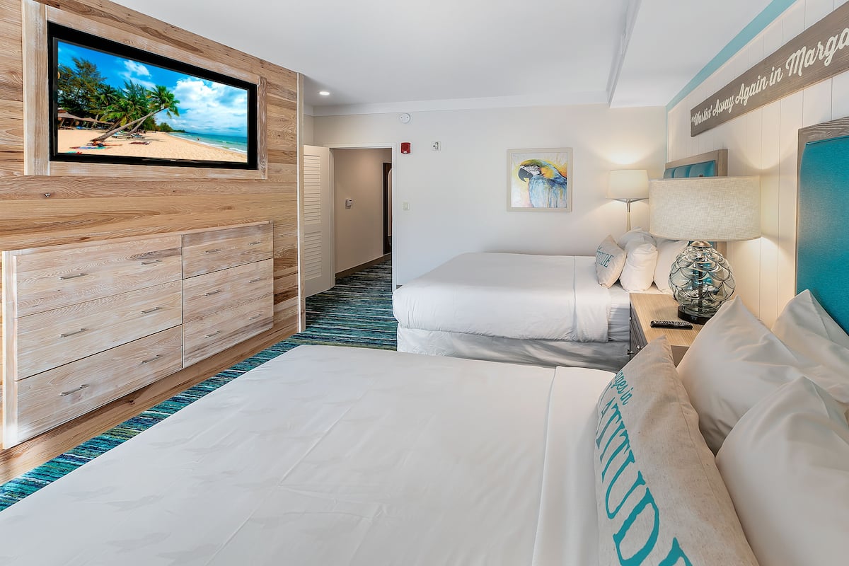 Une chambre d’hôtel avec un grand téléviseur fixé au mur, un lit avec une literie blanche, une commode en bois et un couloir menant à une autre pièce.