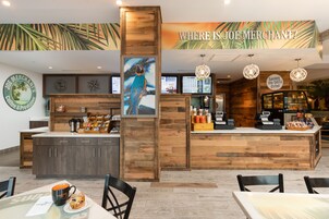 Interior de una cafetería con mostrador de madera, pizarras con el menú y un mural tropical.