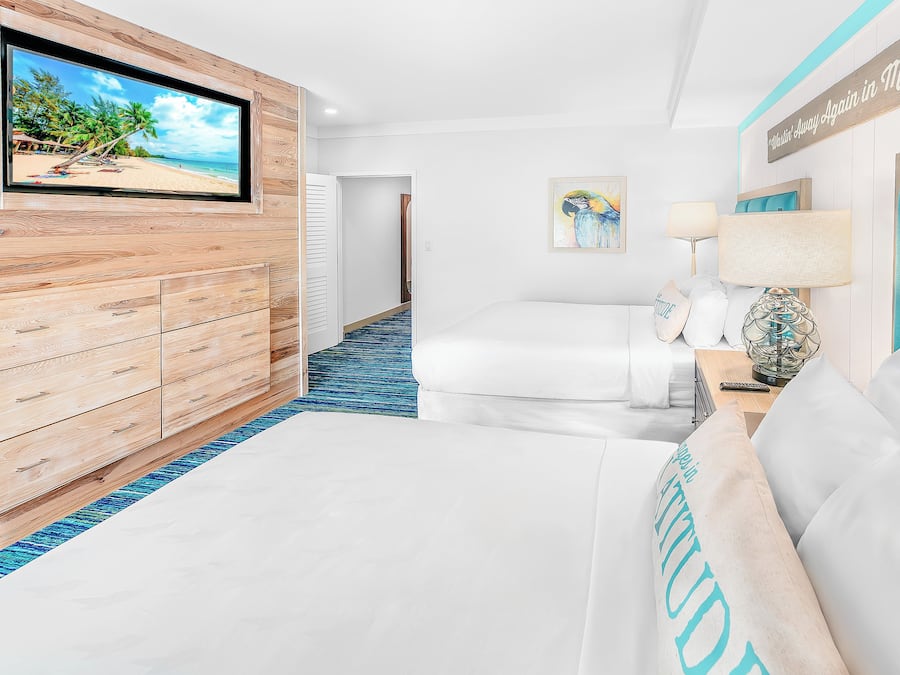 A hotel room with a large TV displaying a beach scene, a wooden dresser, a bed with white bedding, a blue carpet, and a painting of a parrot.