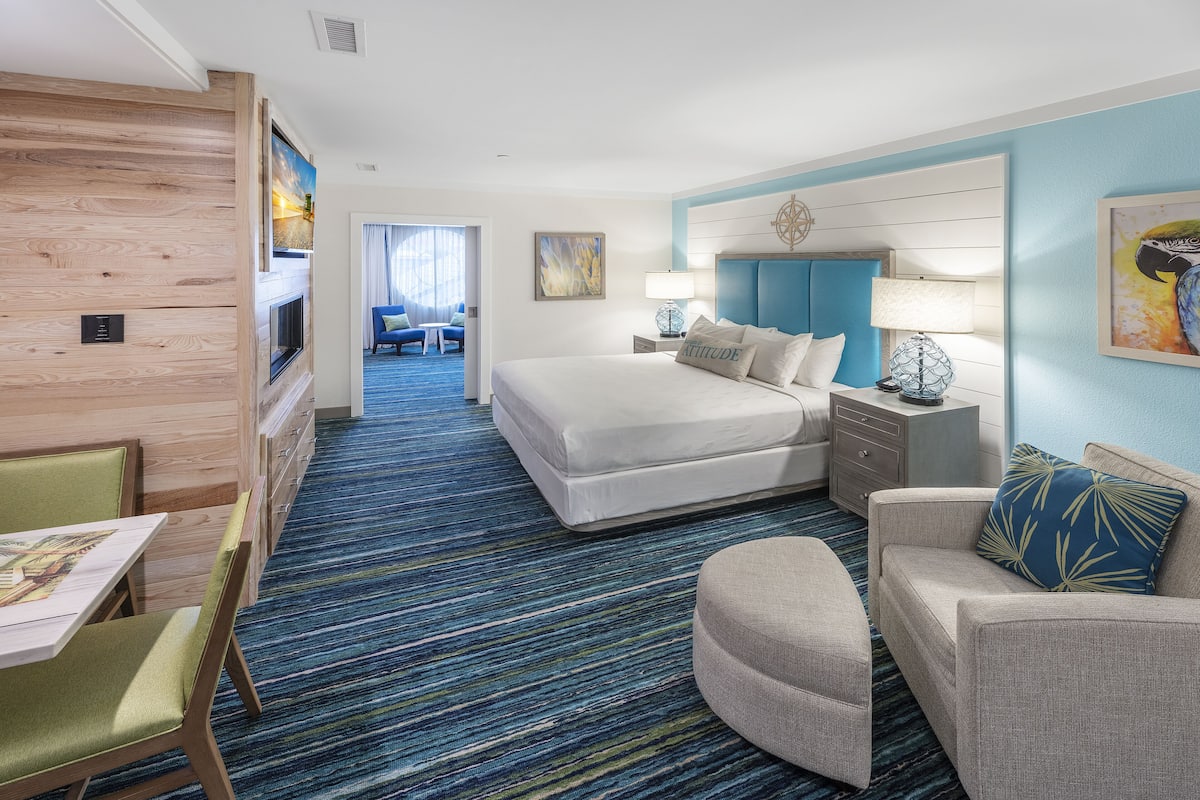 Une chambre d’hôtel avec un grand lit, un coin salon avec une table et des chaises, une télévision et une vue sur le couloir.