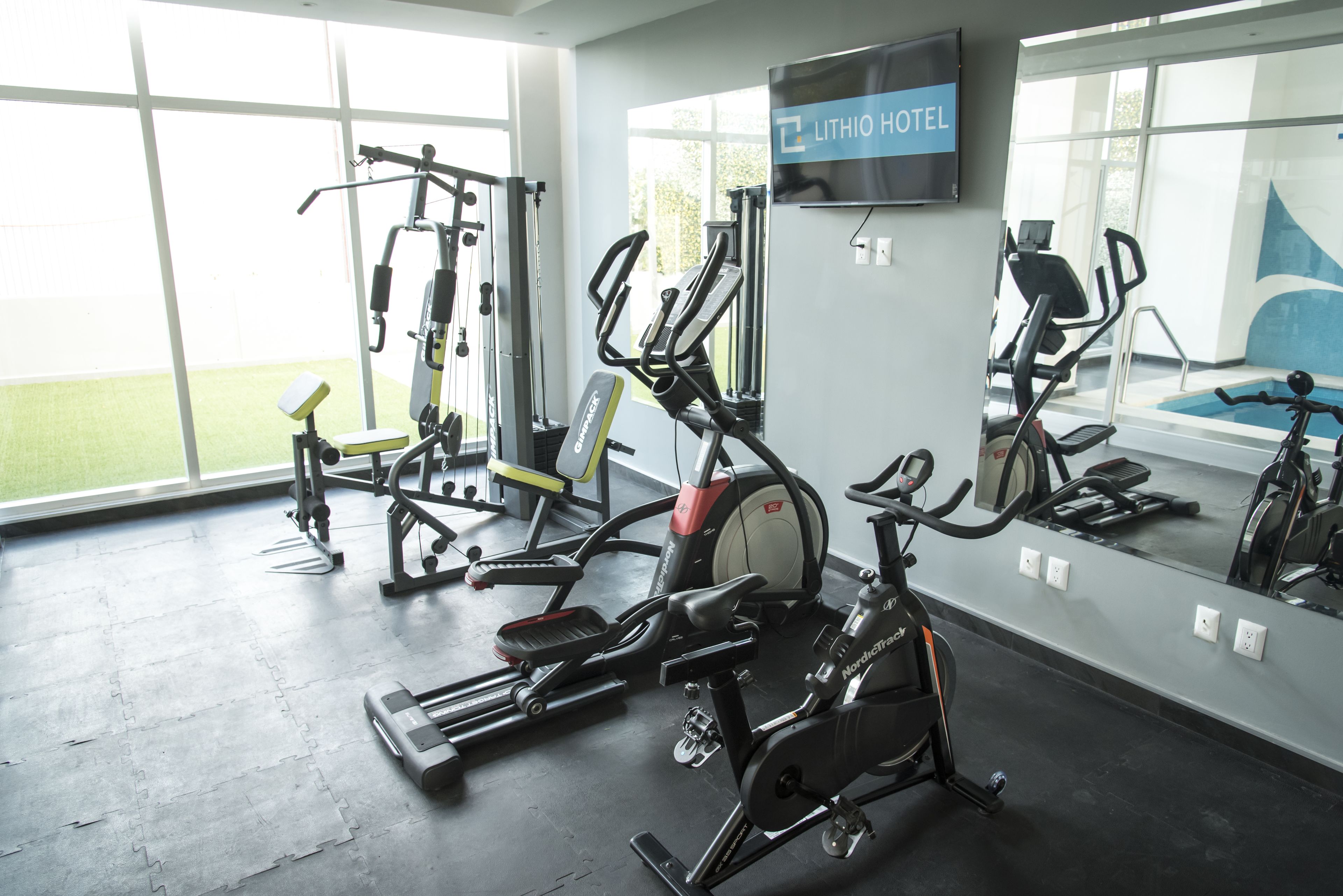 Een fitnessruimte met diverse trainingsapparaten, een groot raam en een bord met de tekst LITHIO HOTEL.