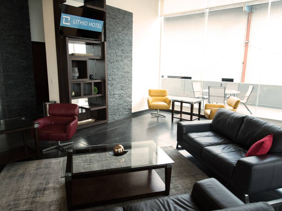 Um hall de hotel moderno com uma mesa de centro de vidro, sofás de couro, uma cadeira vermelha e uma estante de livros.