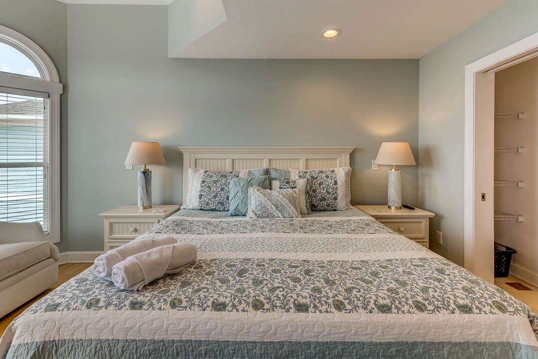 A bedroom with a large bed, two bedside tables with lamps, a window with blinds, and a door to another room.