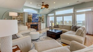 A spacious living room with a fireplace, large windows, and a view of the sea. - Pebble Beach | Oceanfront | Dog Friendly, Private Pool, Hot Tub | Nags Head (Nags Head)