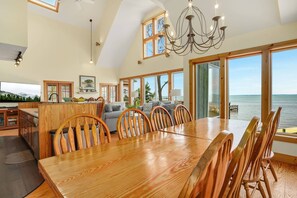 A spacious dining area with a large wooden table and chairs, a kitchen with wooden cabinets, and large windows offering a view of the sea. - Grandview | Luxury Waterfront Sunset Views w/ Long Pier & Dock, Screened Porch, Kayaks + Pets (Chincoteague Island)