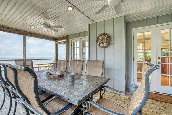 A covered outdoor dining area with a table and chairs, a ceiling fan, and a view of the ocean. - Grandview | Luxury Waterfront Sunset Views w/ Long Pier & Dock, Screened Porch, Kayaks + Pets (Chincoteague Island)