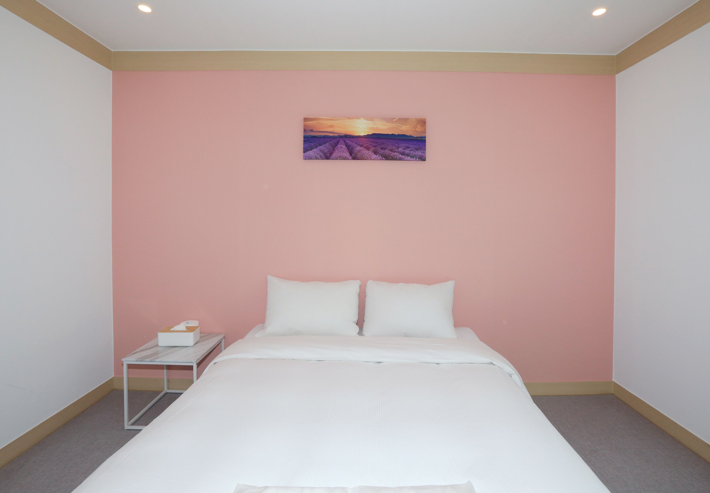 A bedroom with a bed, white bedding, a nightstand with a tissue box, and a painting of a sunset on the wall.