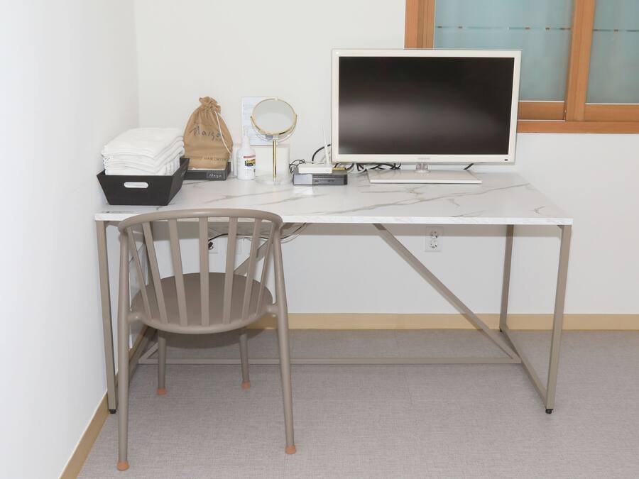 Un escritorio blanco con un monitor de computadora, una silla, un espejo pequeño y una bolsa de productos para el cabello.