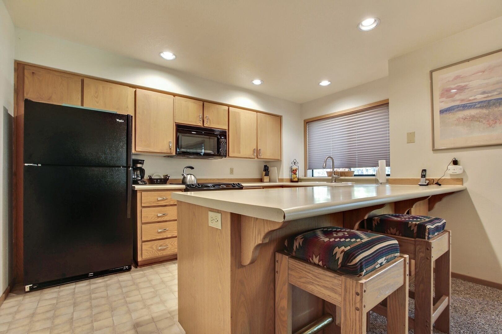 Cocina compacta con armarios de madera, un refrigerador negro, microondas y pileta. Hay dos taburetes en la isla.
