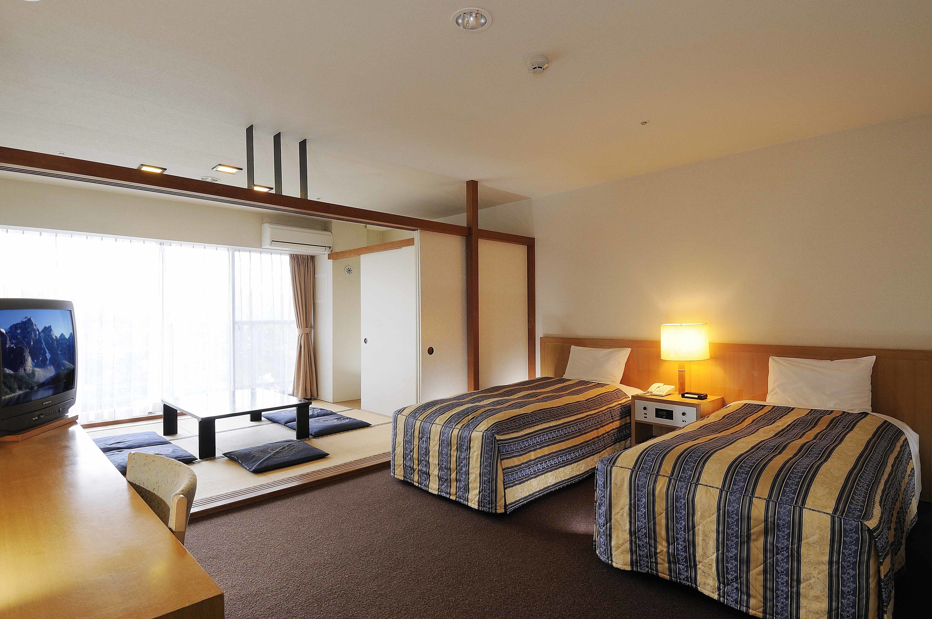 A hotel room with two beds, a desk, a TV, and a window with curtains.