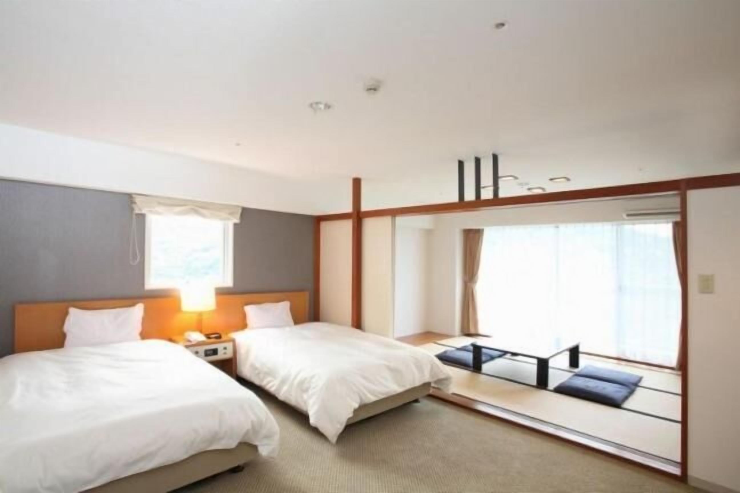 Hotellihuone, jossa on kaksi sänkyä, yöpöytä lampun kanssa, suuri ikkuna verhoilla ja liukuovi, joka johtaa parvekkeelle.