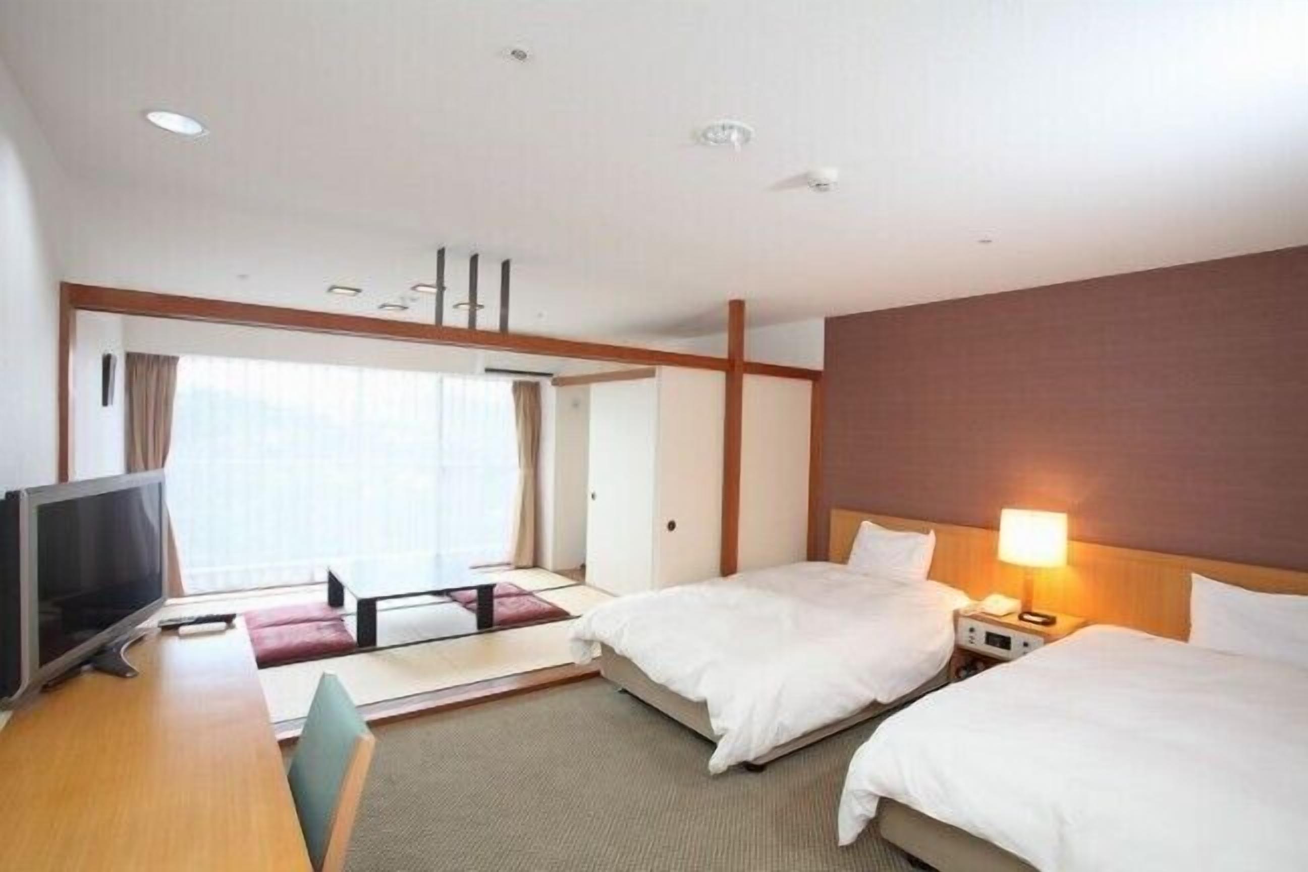 A hotel room with two beds, a desk with a TV, and a large window.