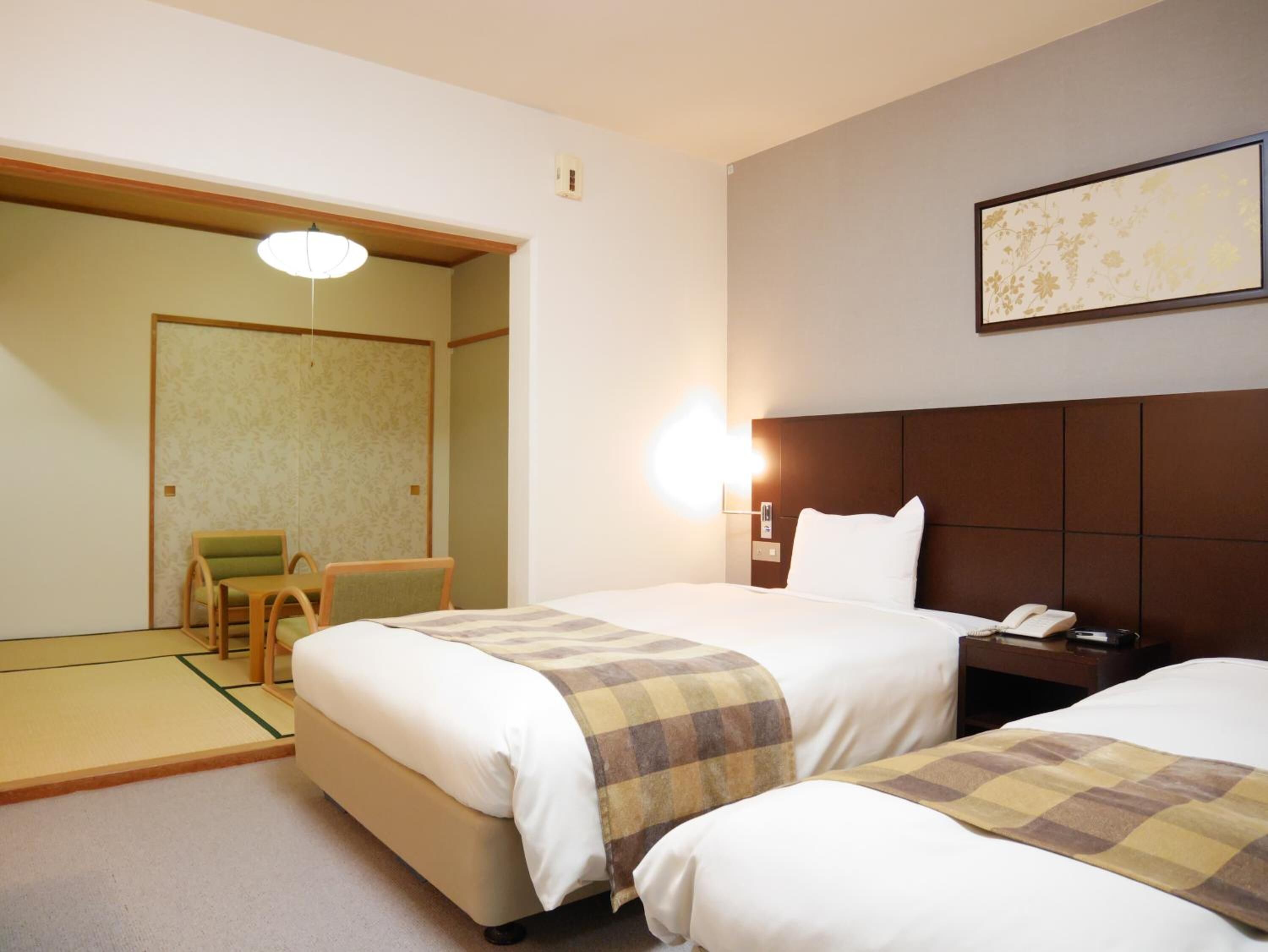 A hotel room with two beds, a chair, a bedside table with a phone, and a wall-mounted light.