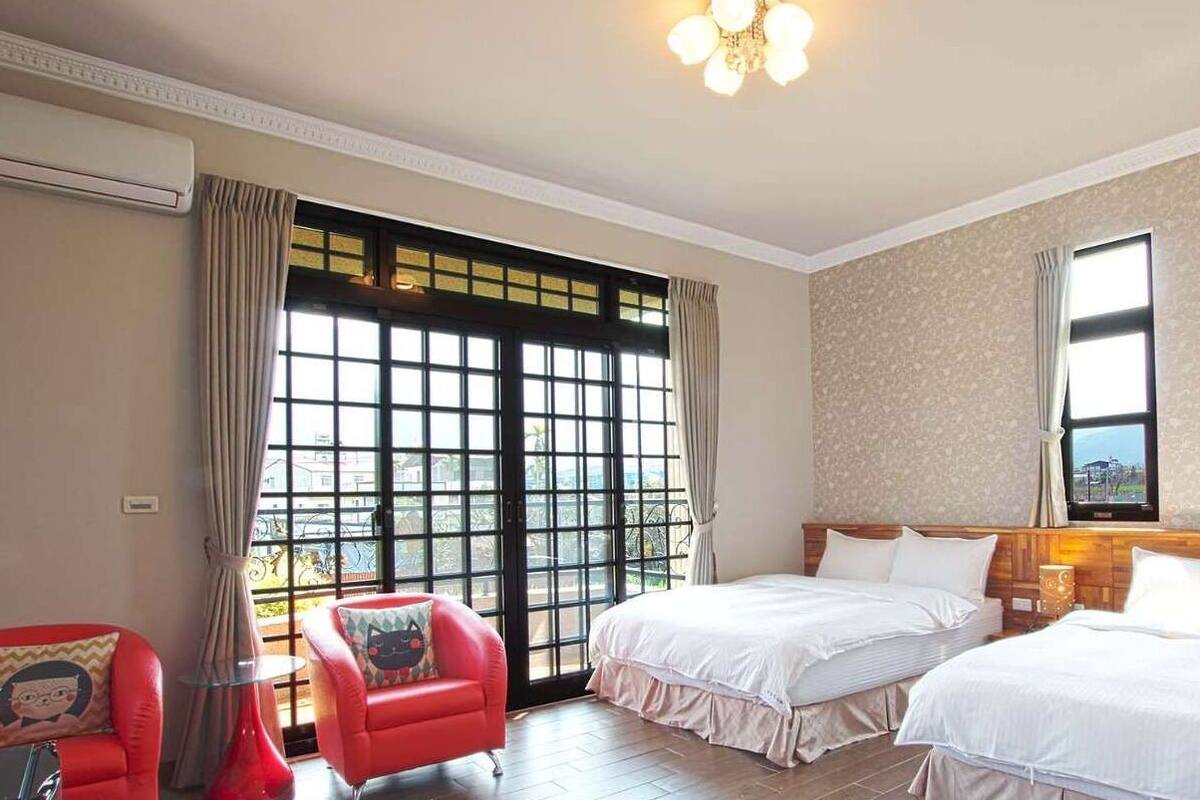 A hotel room with two beds, a red armchair, a small table, and a large window with curtains.