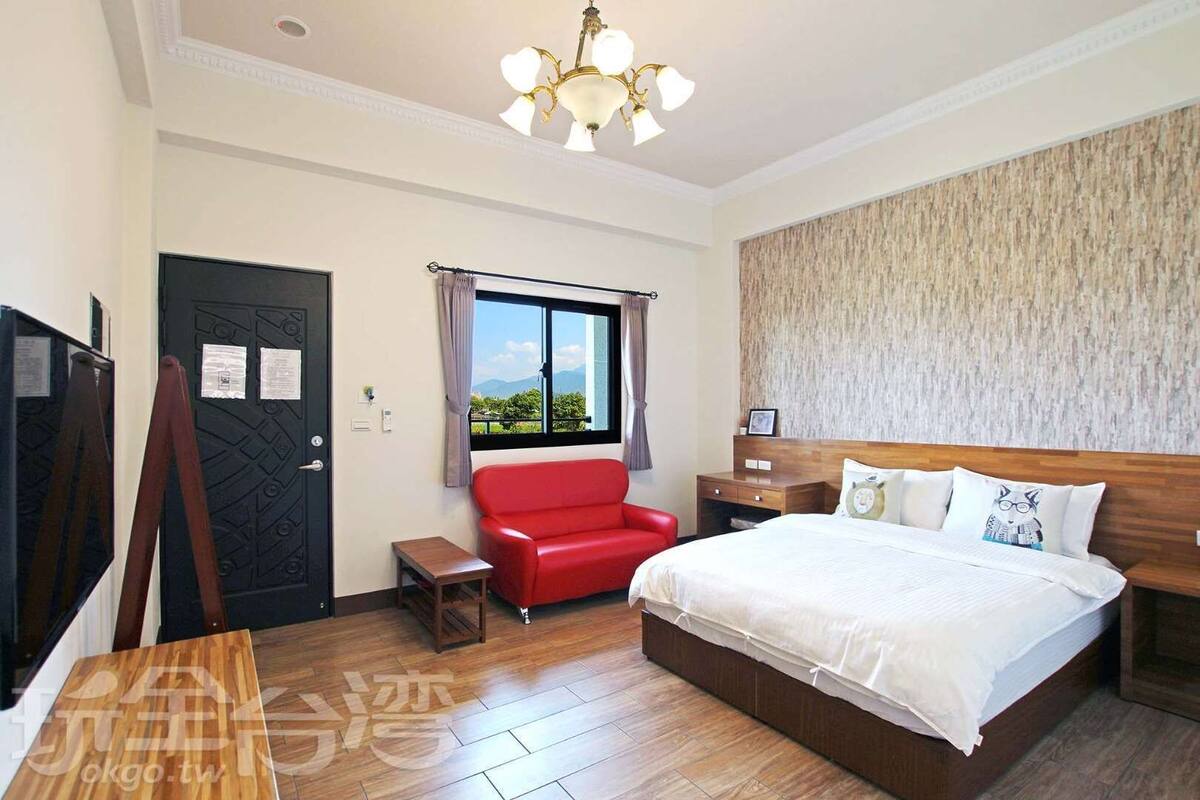 A hotel room with a large bed, a red armchair, a small table, a TV, and a window with a view.