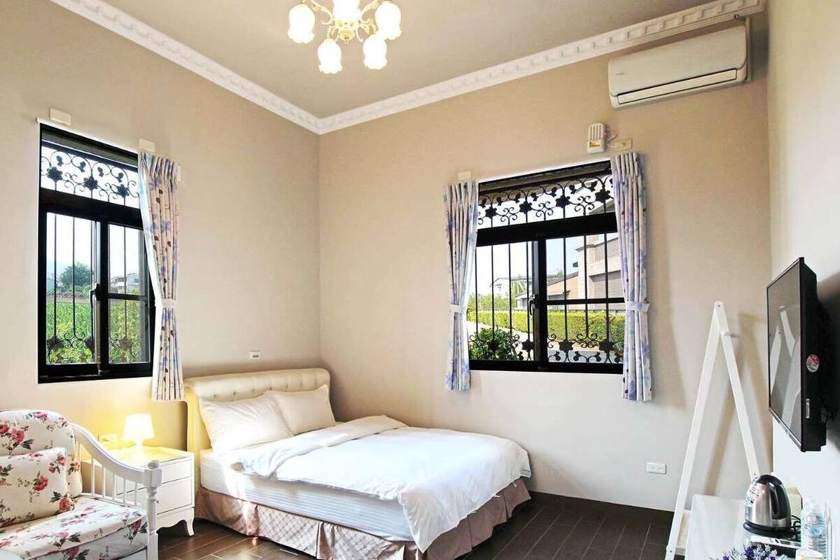 A bedroom with a bed, a chair, a nightstand, a TV, and two windows with curtains.