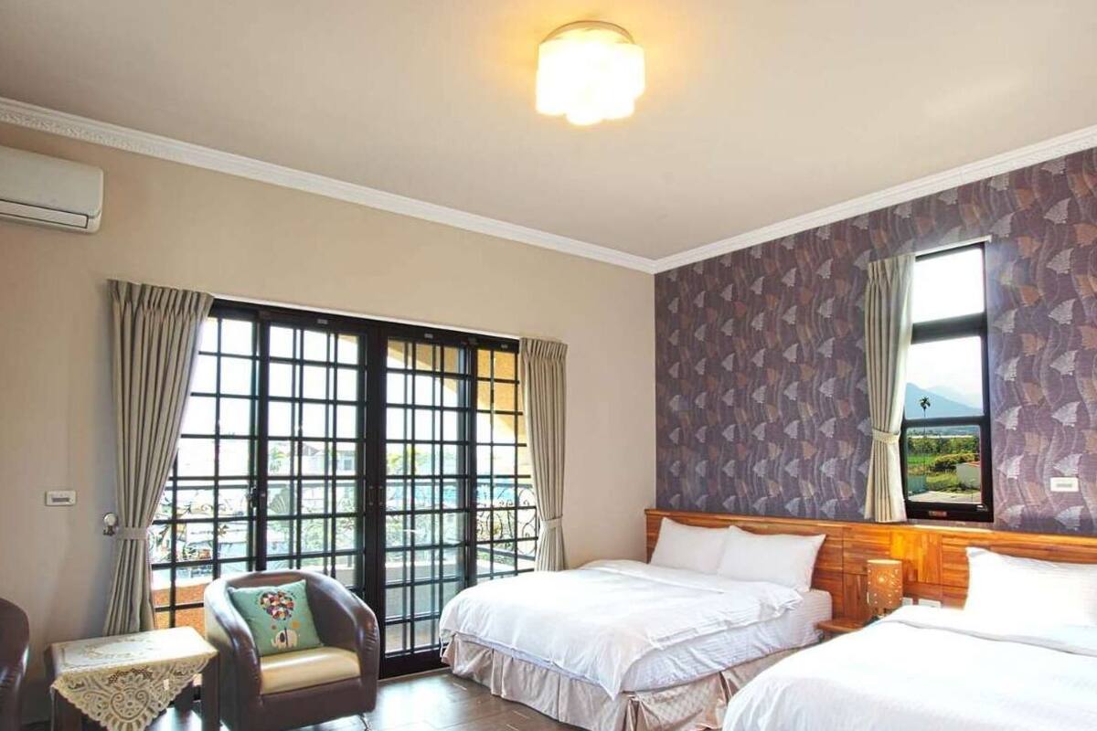 A hotel room with two beds, a large window with curtains, a chair, and a table.