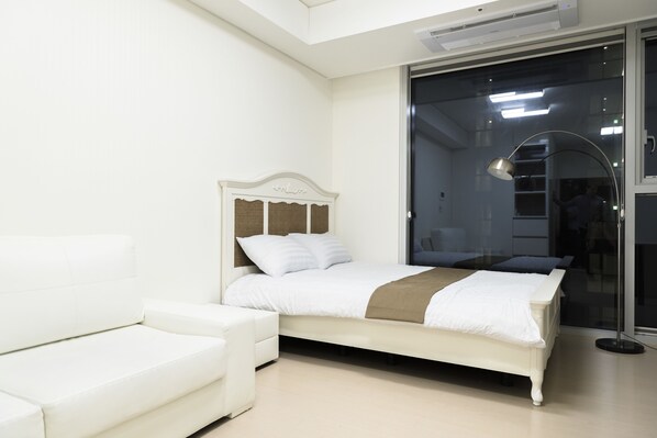 A modern bedroom with a white bed, a white sofa, and a large window with a view of the city.