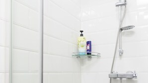 A modern bathroom with white tiled walls, a shower with a handheld showerhead, and two glass shelves with toiletries.