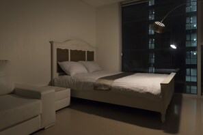 A bedroom with a bed, a nightstand, a lamp, and a window with a view of buildings.