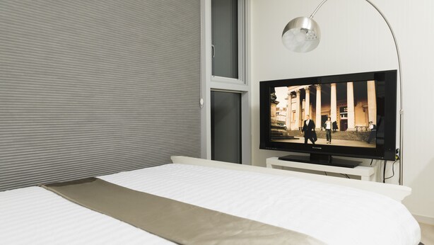A modern hotel room with a large bed, a flat-screen TV mounted on a white stand, and a large window with blinds.