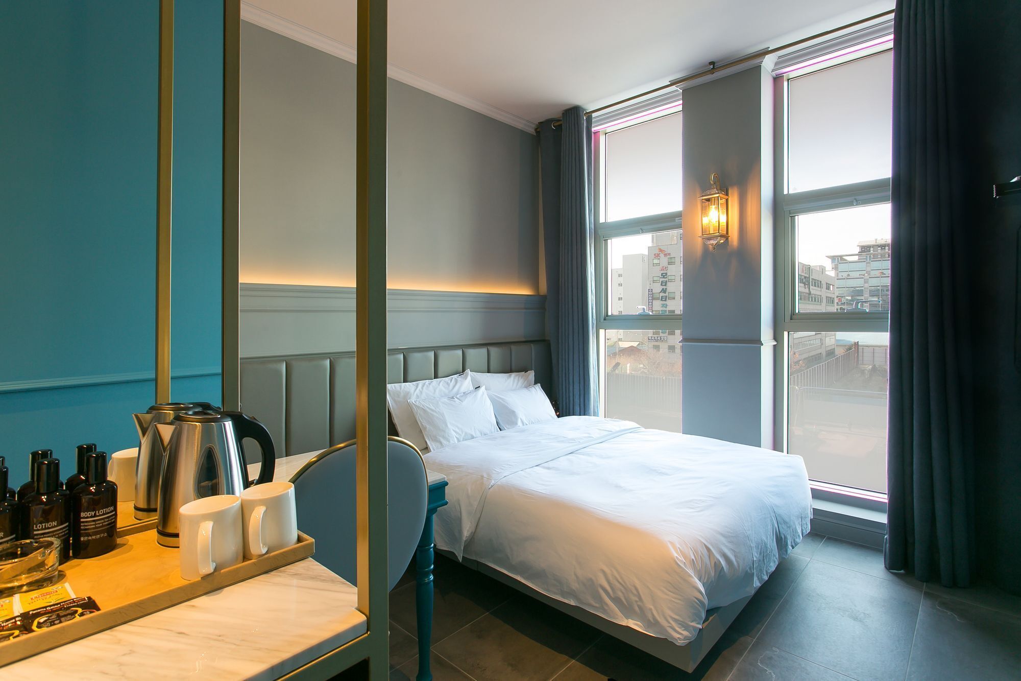 A hotel room with a bed, a bedside table with toiletries, a chair, and a large window with a city view.