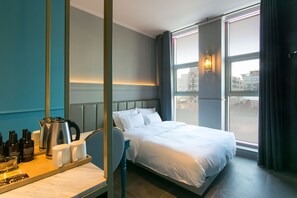 Habitación de hotel con cama, mesita de noche con artículos de aseo, una silla y un ventanal con vistas a la ciudad.