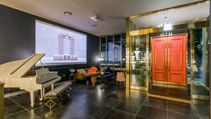 Eine moderne Hotelhalle mit einem Flügel, bequemen Sitzgelegenheiten, einem großen Bildschirm, der eine Stadtansicht zeigt, und einer auffälligen, roten Tür.