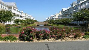 A row of white, multi-story buildings with balconies, a stone bridge, and a landscaped area with pink flowers.