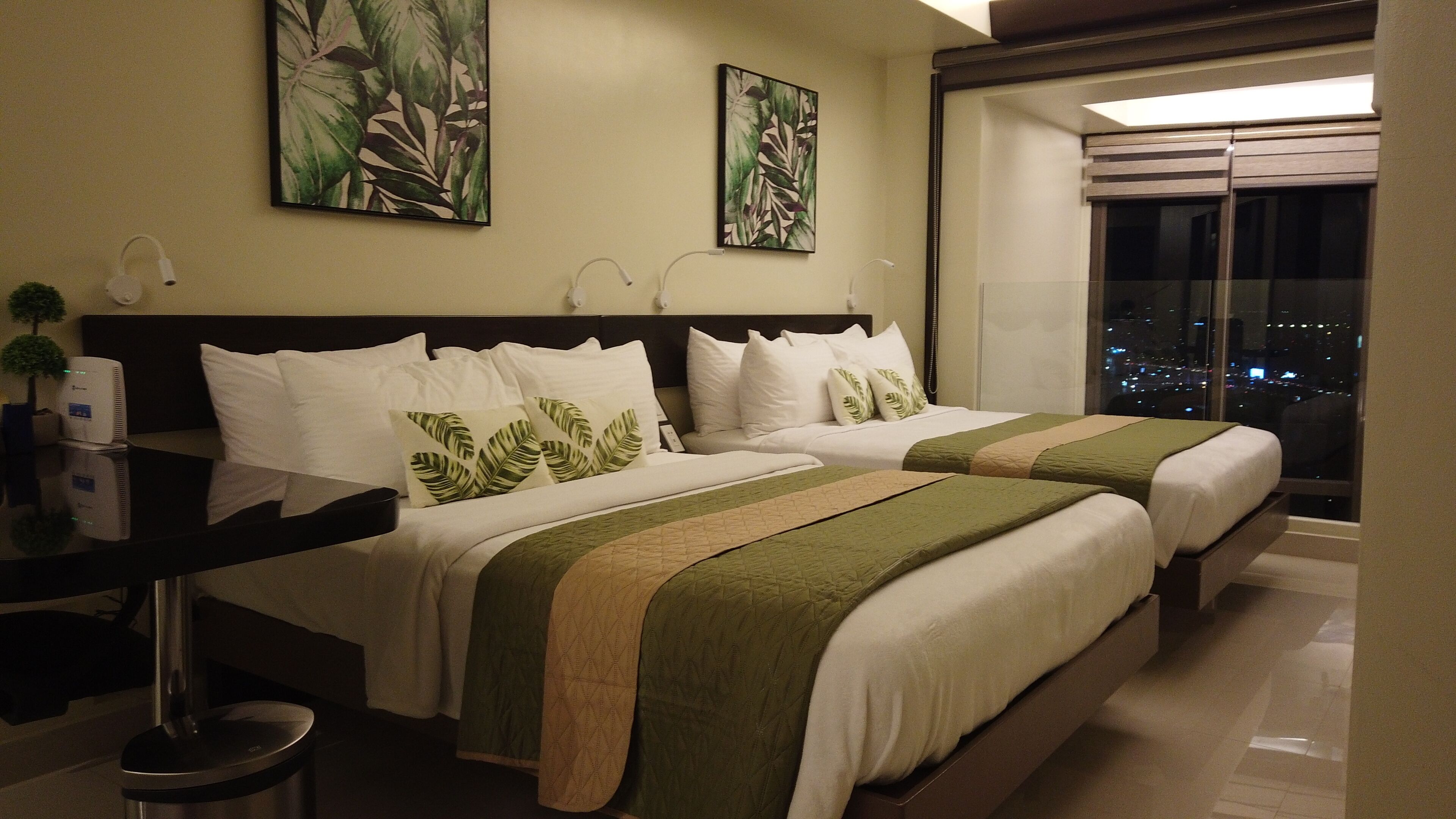A hotel room with two beds, a nightstand, a desk, and a view of the city at night.