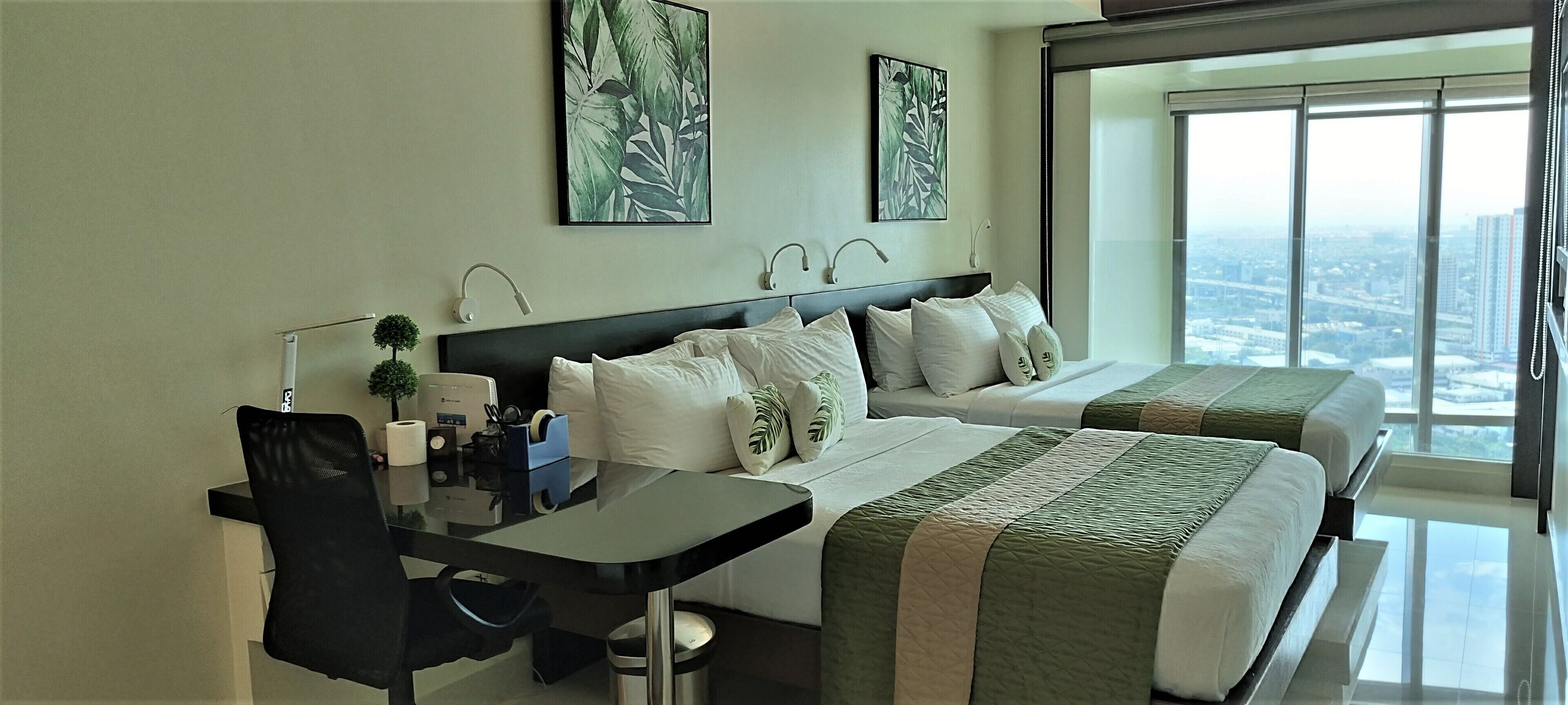 A hotel room with two beds, a desk, and a large window offering a city view.