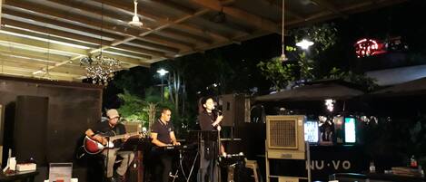 Eine Band tritt auf einer Bühne mit Mikrofonstativ und Musikinstrumenten auf. Im Vordergrund stehen Tische und Stühle, und es gibt eine Bar mit Theke und Hockern.