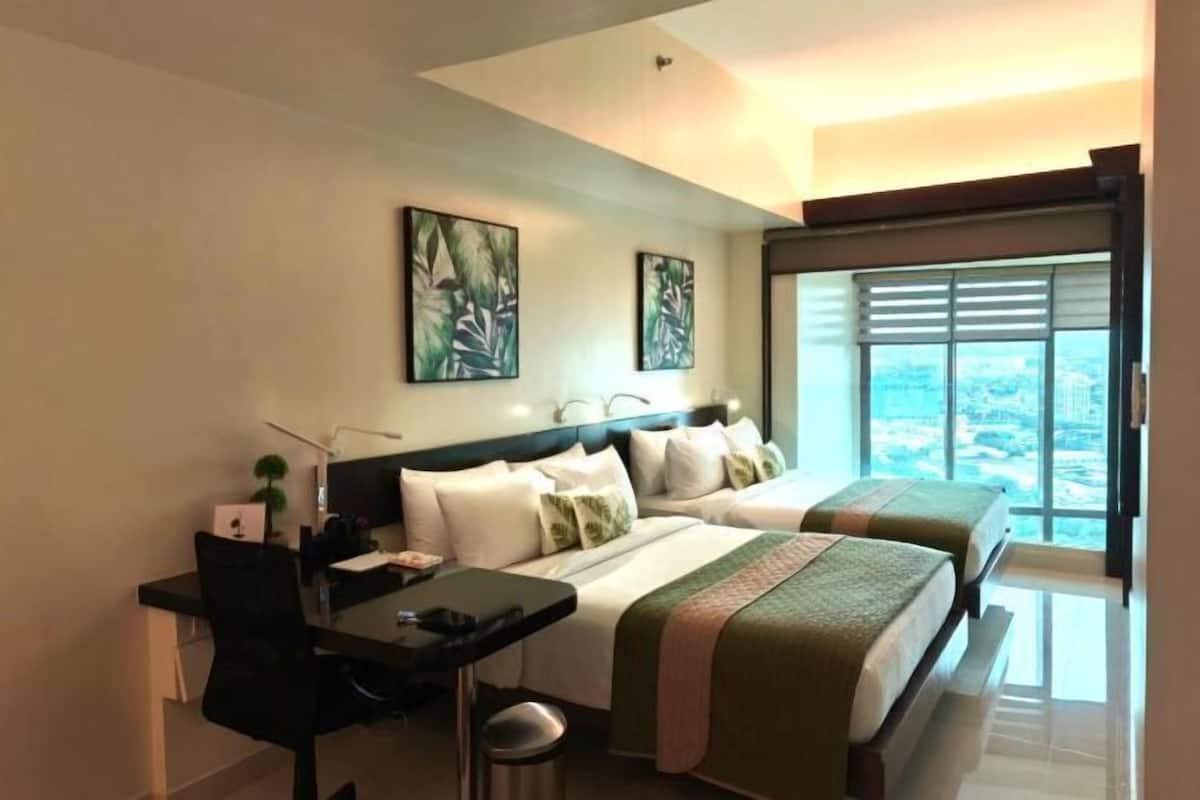 Quarto de hotel com duas camas, uma mesa, uma cadeira e uma janela grande com vista para a cidade.