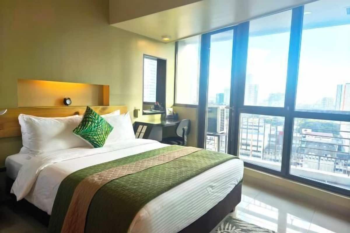 Quarto de hotel com uma cama grande, uma mesa, uma cadeira e vista da cidade através de grandes janelas.