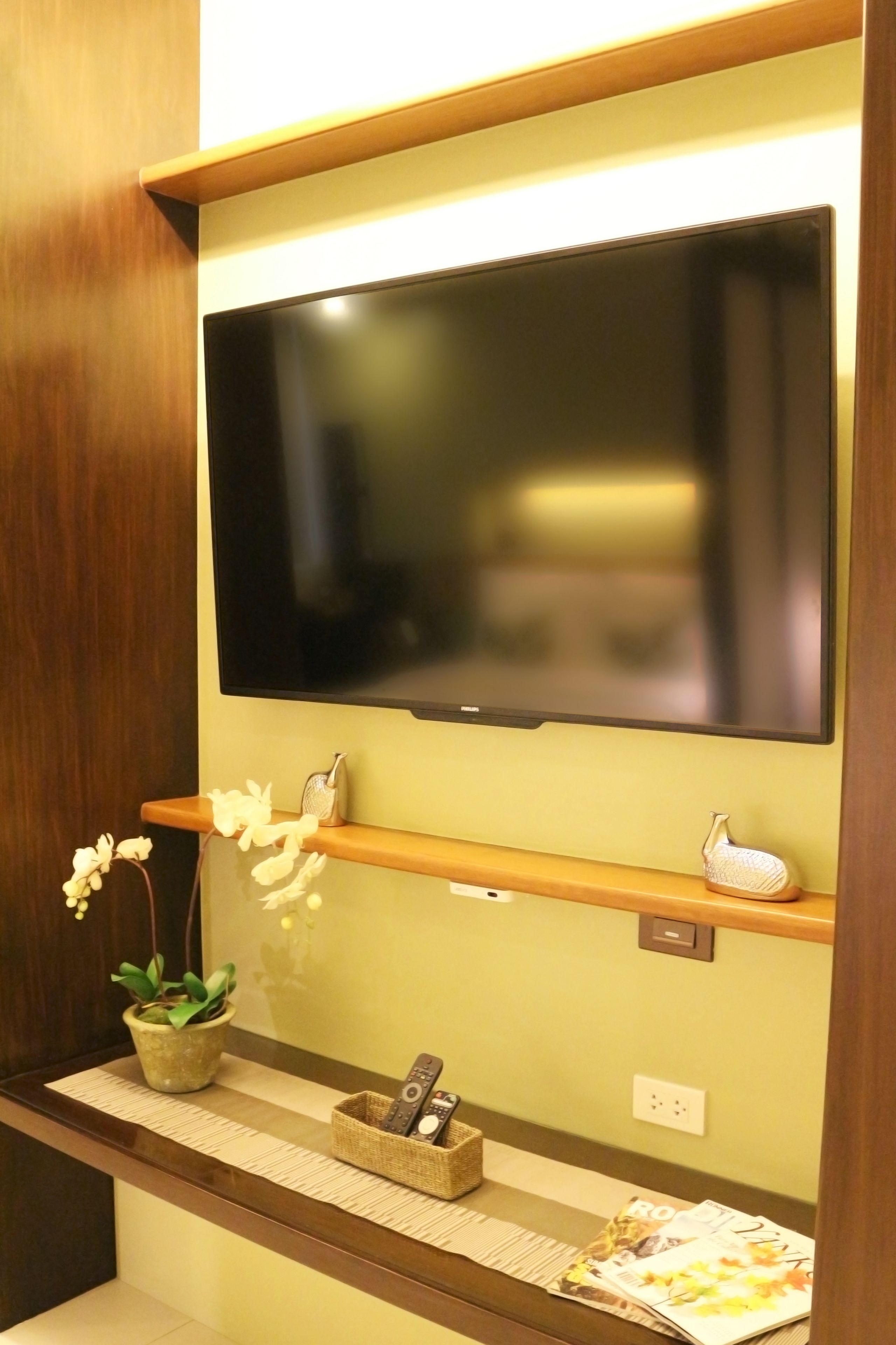 A television mounted on a wall with a wooden shelf below. There is a potted plant on the left and a basket with remote controls on the right.